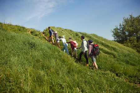 Group of young people, friends walking up the hill on warm spring day. Men and women going hiking,...