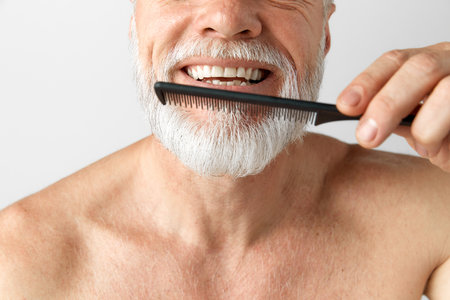 Portrait of handsome mature man with gray hair taking care after beard, brushing it, posing shirtless against grey backgroundの写真素材