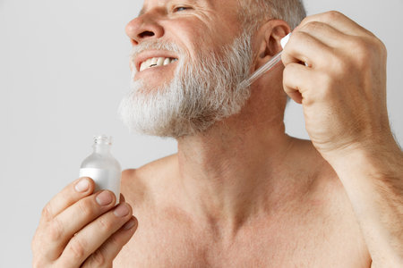 Cropped image of smiling, handsome mature man posing shirtless, applying face serum. Model against grey backgroundの写真素材