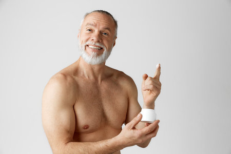 Portrait of positive, smiling, handsome, mature, gray-haired man applying face moisturizing cream against grey backgroundの写真素材