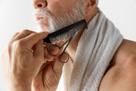 Home barber care. Cropped image of mature mans face, male model taking care after face, cutting and brushing beard against grey backgroundの写真素材