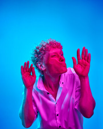 Portrait of man with curly hair, in white shirt, leaning on transparent glass against blue studio background in pink neon light. Funny faceの写真素材