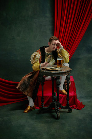 Young sad man sitting at table and drinking lager beer with fish against dark green, vintage backgroundの写真素材