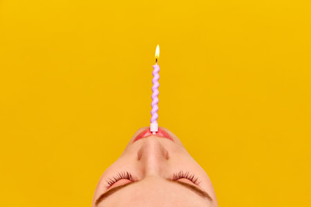 Cropped part of female face with burning candle against vivid yellow background. Making birthday wishesの写真素材