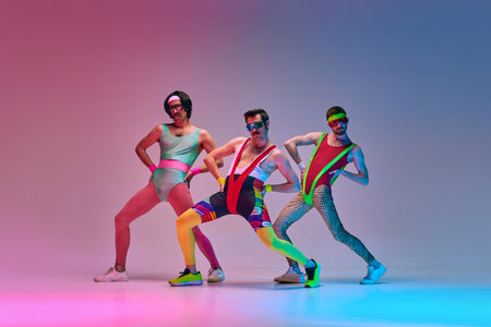 Funny men in vintage, colorful sportswear posing, doing aerobics exercises against gradient blue pink studio background in neon lightの写真素材