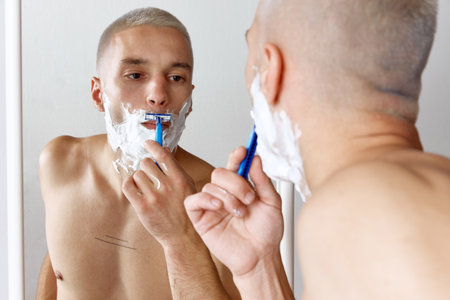 Portrait of young shirtless man shaving his beard, looking in mirror against white studio backgroundの写真素材