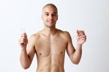 Portrait of young, shirtless, handsome man taking care after skin with cosmetics, face serum against white studio backgroundの写真素材