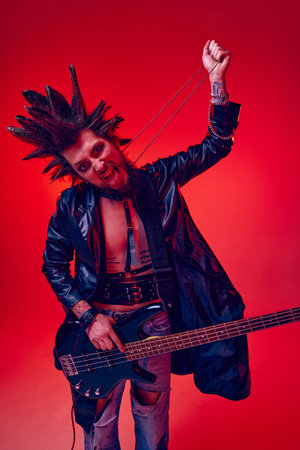 Young man, punk in extraordinary clothes and hairdo playing electric guitar against red studio background in neon light. Crazinessの写真素材