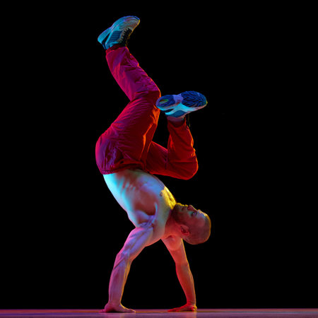 Young talented, expressive man with muscular body dancing breakdance, hip-hop against black studio...