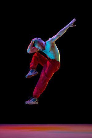 Young muscular man with relief shirtless body dancing breakdance against black studio background in neon lightの写真素材