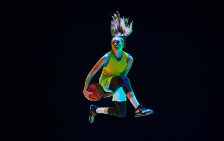 Dynamic image of motivated sportsman, young girl playing basketball, jumping against black studio background in neon lightの写真素材