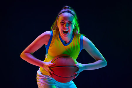 Concentrated and motivated young girl, basketball player in uniform posing with ball against black studio background in neon lightの写真素材