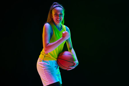 Winner. Young girl, basketball player in uniform posing with ball against black studio background in neon lightの写真素材