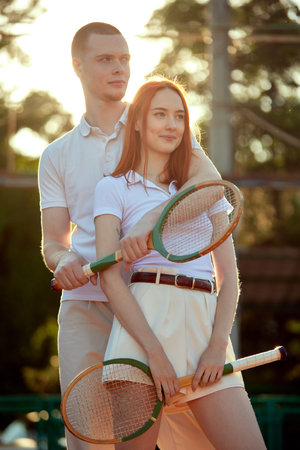 Beautiful young redhead man and woman in stylish, casual, light clothes posing with tennis racket on warm day during sunset timeの写真素材