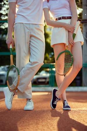 Cropped image of man and woman in stylish, light, casual clothes posing with tennis racket on open air court during sunsetの写真素材