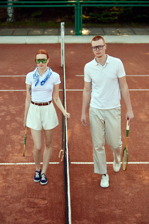 Full-length image of beautiful young woman and handsome man in stylish clothes and stylish eyewear posing on open air tennis court on daytimeの写真素材
