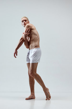 Full-length portrait of handsome young man with blonde hair, muscular body posing in underwear against grey studio backgroundの写真素材