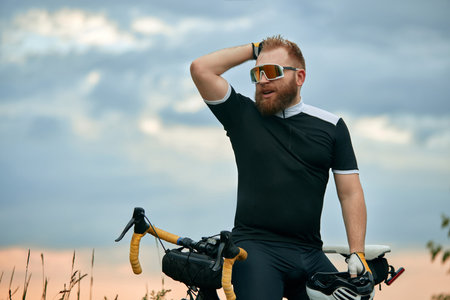 Handsome, young, bearded man in sport glasses and sportswear, cyclist sitting in bike over field backgroundの写真素材