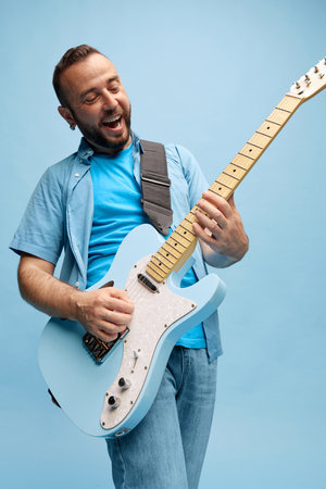 Smiling bearded man in casual clothes emotionally playing guitar against blue studio background. Lifestyleの写真素材