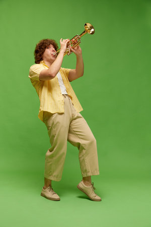 Full-length portrait of young man with curly hair, musician playing trumpet against green studio backgroundの写真素材