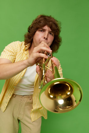 Portrait of young man with curly hair, talented musician playing trumpet against green studio...