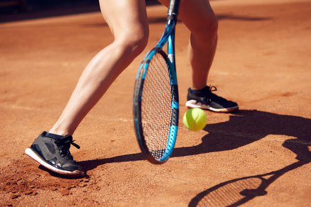 Cropped image of female legs in sportswear and tennis racket with ball over floor background with shadows. Open air tennis courtの写真素材