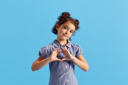 Portrait of lovely little girl, child with curly hair and cute hairdo posing with heart shape with hands against blue studio backgroundの写真素材
