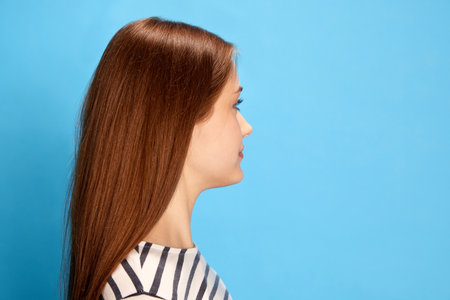 Side view portrait of young girl with straight hair looking forward, posing against blue studio backgroundの写真素材