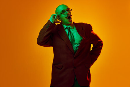 Portrait of young, emotive, bald man with moustache standing with excited thoughtful face against orange studio background in neon lightの写真素材