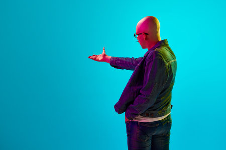 Side view portrait of young bald man in casual clothes showing away against blue studio background in neon lightの写真素材