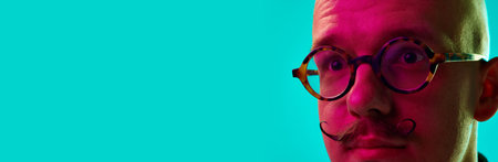 Close-up cropped image of male face, young bald man with moustache in glasses against blue studio background in neon lightの写真素材