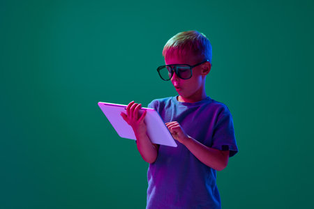 Online games. Little boy, child in sunglasses playing on tablet with excitement against cyan studio background in neon lightの写真素材