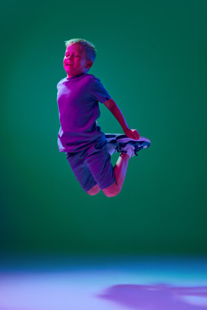 Playful little boy, child in casual clothes cheerfully jumping against cyan studio background in...