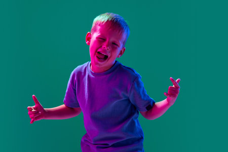 Portrait of little playful boy, child in casual clothes playing, showing funny face against cyan studio background in neon lightの写真素材