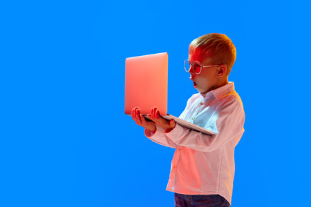 Online lessons. Emotional boy, child in glasses looking with shocked face in laptop against blue studio background in neon lightの写真素材