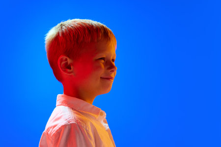 Portrait of positive little boy child in white shirt design with smile against blue studio background in neon lightの写真素材