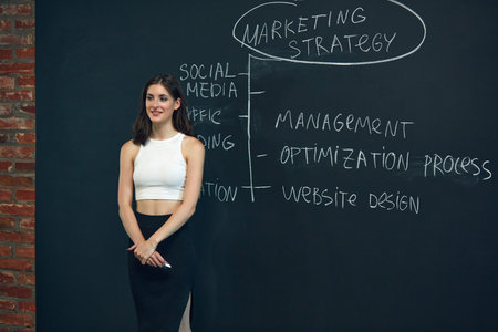 Young woman, marketing department employee representing marketing plan on blackboard. Conference and...