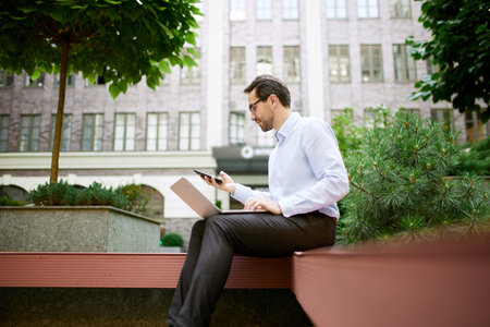 Young businessman sitting on bench outside of his office and working online with laptop and mobile...