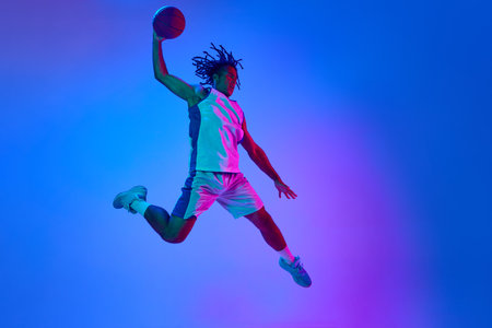 Competitive, ambitious sportsman, young basketball player in motion with ball against gradient blue background in neon lightsの写真素材