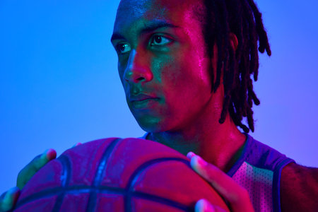 Portrait of handsome young man, basketball player posing with ball against gradient blue background in neon lightsの写真素材