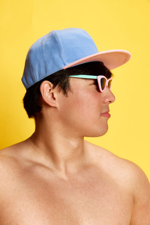 Profile portrait of young man in sunglasses and cap posing shirtless against yellow studio background. Tanned bodyの写真素材