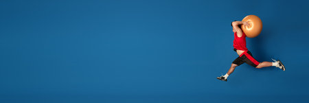 Full-length portrait of young ma in sportswear training, jumping with fitness ball against blue studio backgroundの写真素材