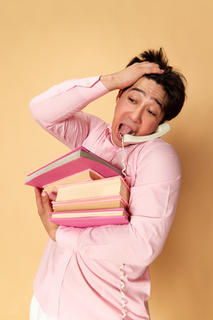 Portrait of young emotional man talking on phone, carrying many books against yellow studio background. Deadlinesの写真素材