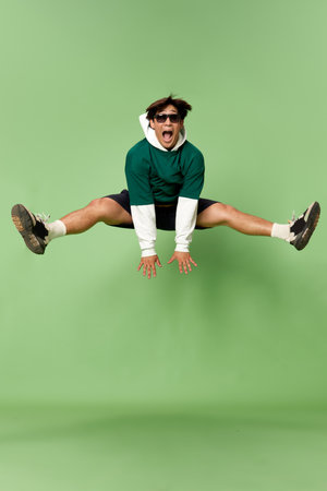 Portrait of young stylish man in sunglasses and sport style clothes cheerfully jumping against green...