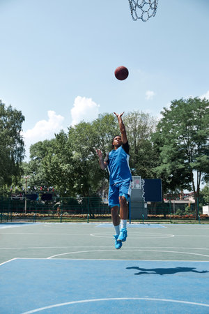 Full-length image of young sportive guy in blue uniform training, playing basketball on...
