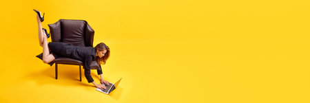 Concentrated business woman lying on armchair and typing on laptop on floor against bright yellow...