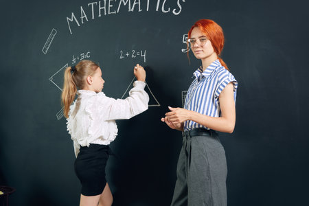 Young woman, teacher of math standing by blackboard with little girl student and solving geometry tasks. Drawingsの写真素材