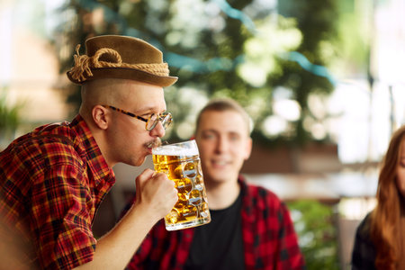 Close-up of young man in checkered shirt and fedora hat drinking beer. Blurred people in background. Sunny dayの写真素材