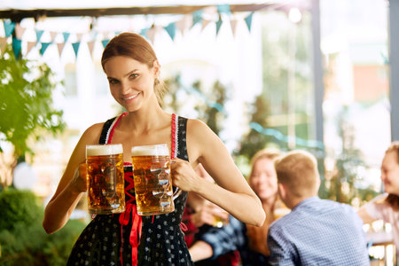 Portrait of beautiful young woman, waitress in bavarian traditional clothes standing with beer glasses. Blurred people on backgroundの写真素材
