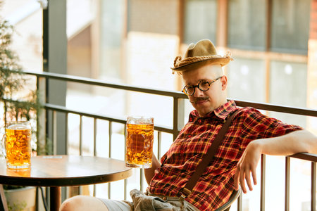 Young stylish man with moustache, in checkered shirt and fedora hat sitting at pub and drinking lager beerの写真素材